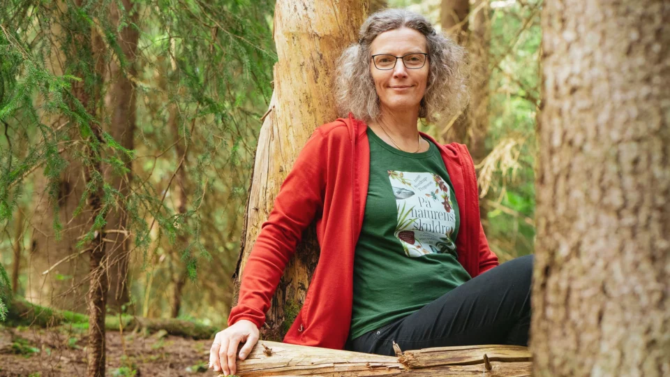 Anne Sverdrup-Thygeson sitter på en trestamme inne i skogen, mellom to store trær, og smiler mot kamera.